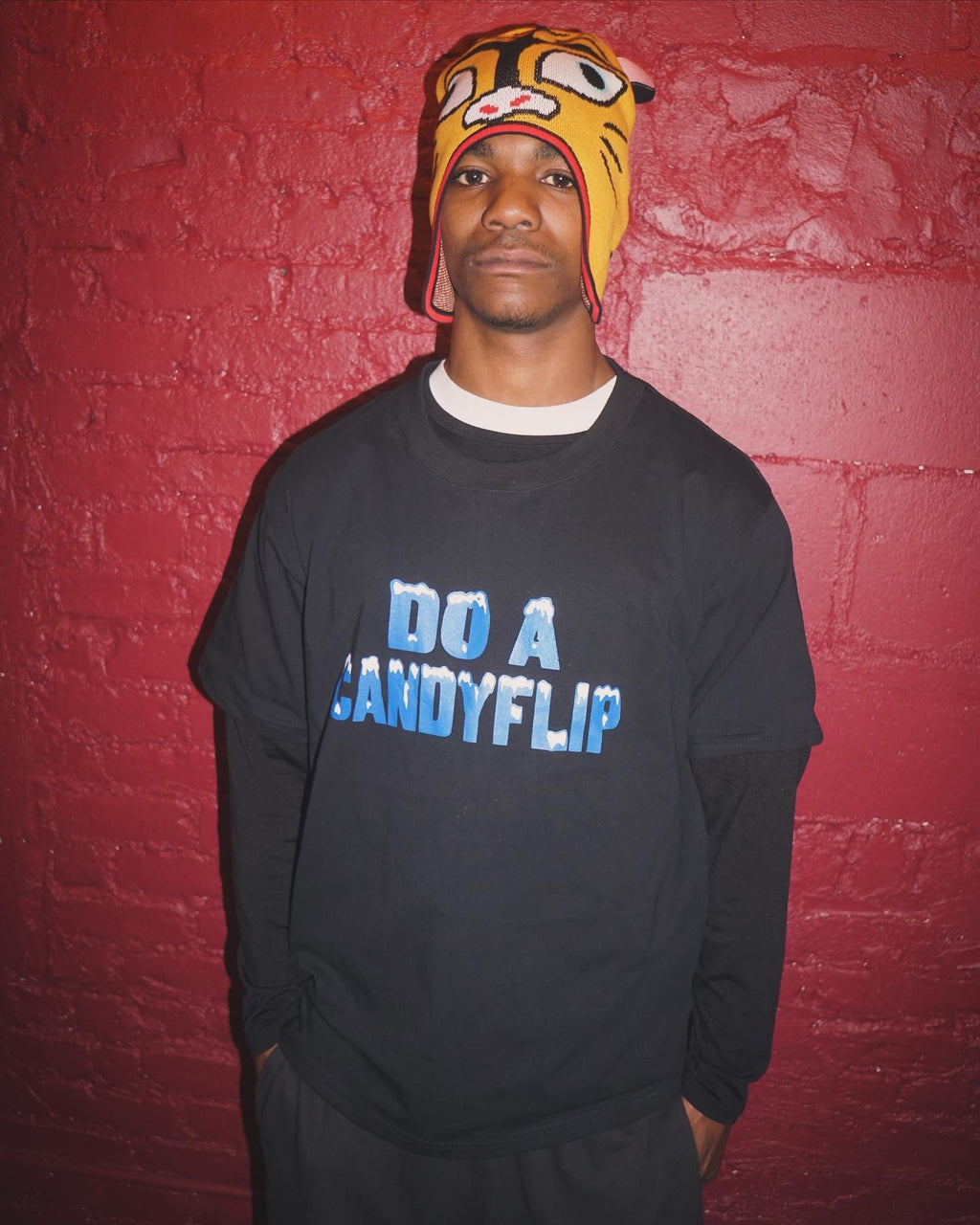 Skateboarder Chucky Lane wearing the Stingwater "Do a Candyflip" T-shirt in black with bold blue font and white icy snow capped letters with a red brick backdrop in NYC